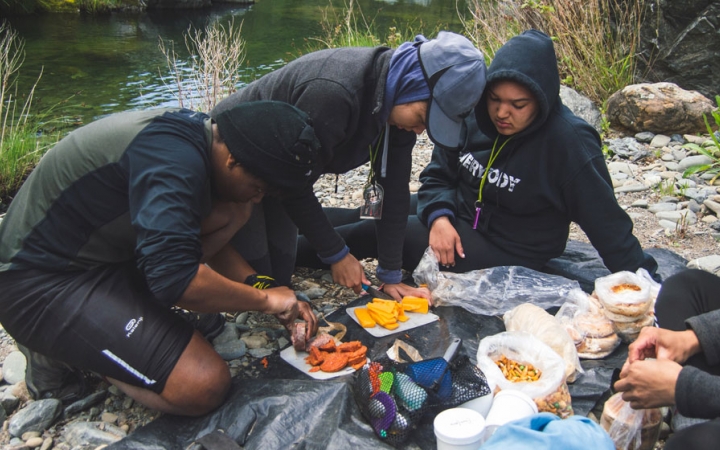 a group of people prepare a meal on a shore beside a body of water on an outward bound course for bipoc